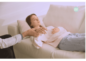 Femme allongée sur un canapé en séance d’hypnothérapie, les yeux fermés, tenue par le poignet par le praticien